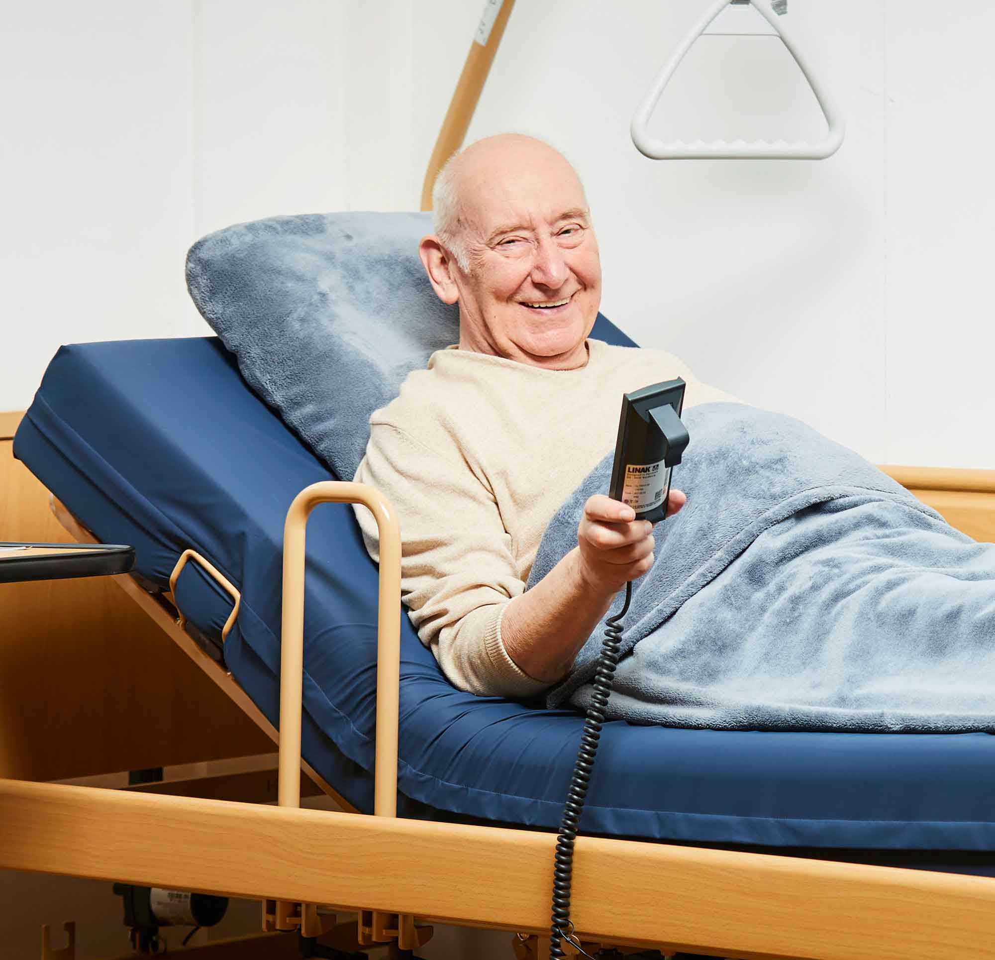 Lächelnder Mann sitzt aufrecht im Pflegebett mit der Bedienungshilfe in der Hand.