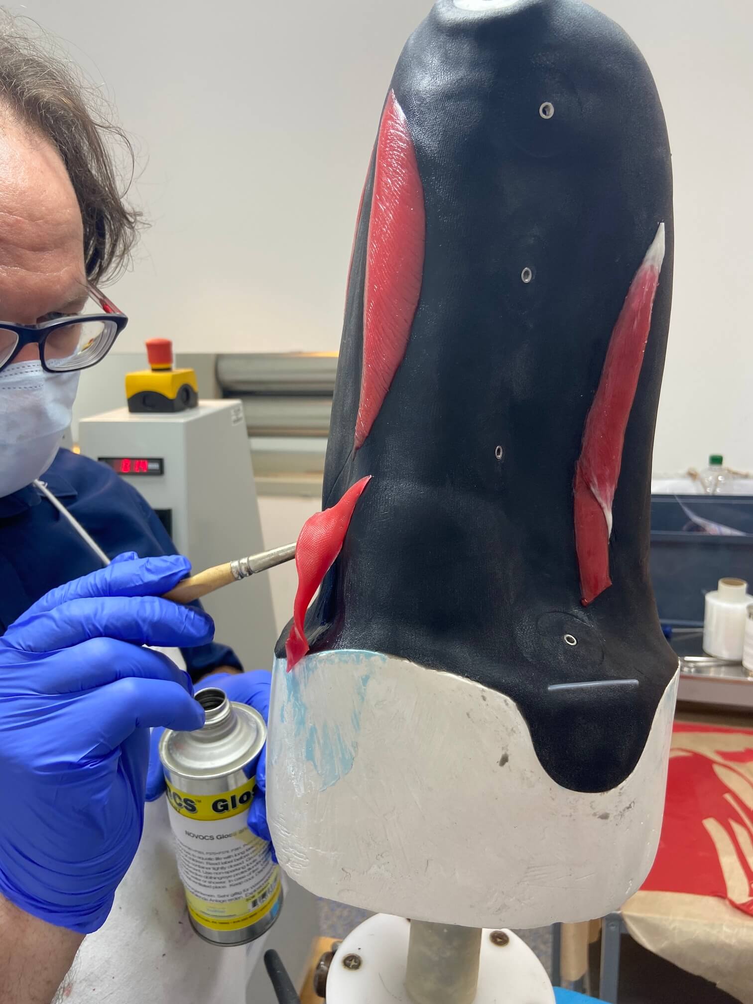 Farbgebung Silikon Herstellungsprozess Ein Orthopädietechniker mit Mundschutz und Handschuhen appliziert präzise rote Silikonelemente, die Muskeln darstellen, auf eine schwarze Prothesenhülle, um eine naturgetreue Ästhetik zu erzielen.