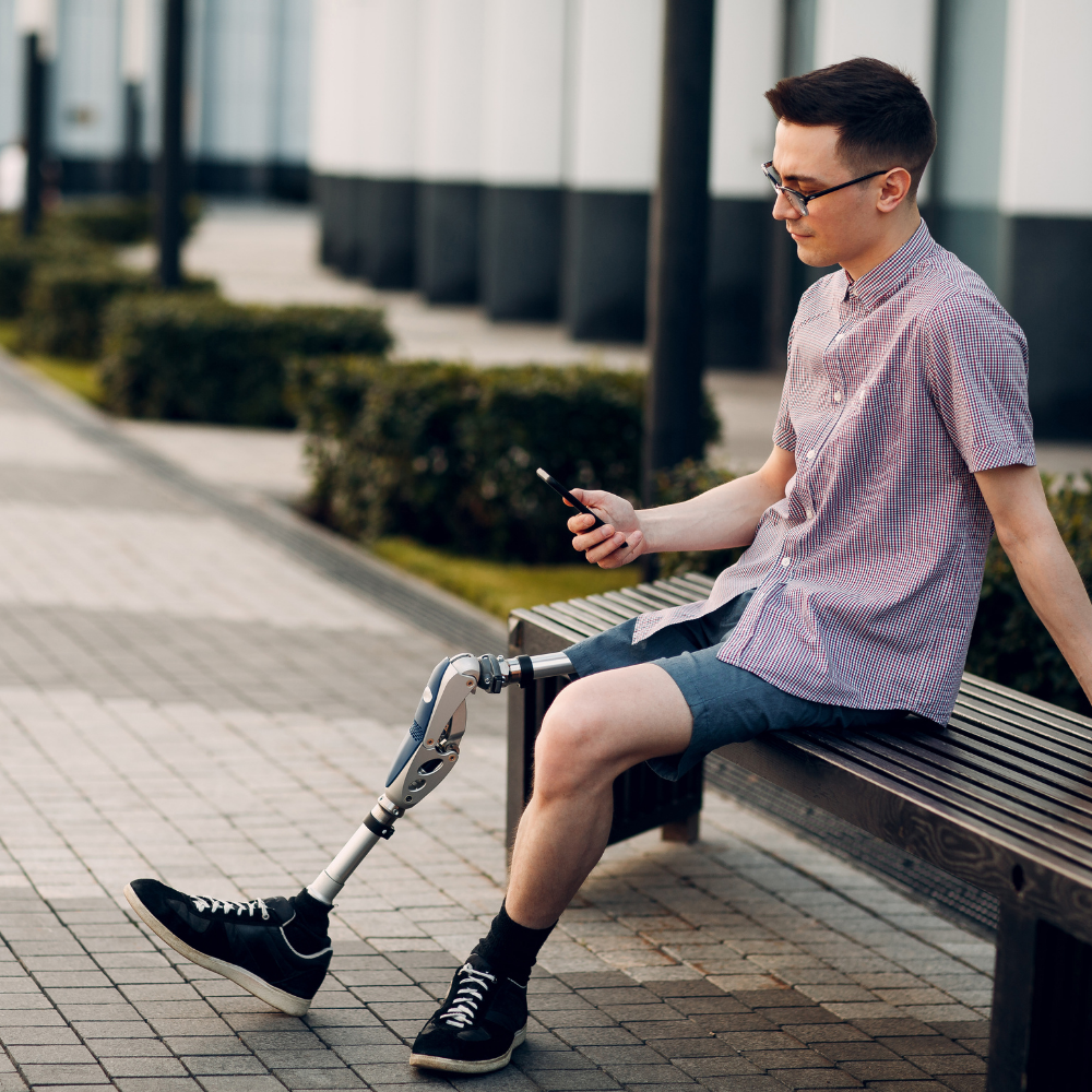 Mann mit Beinprothese sitzt auf einer Bank und schau auf's Handy.