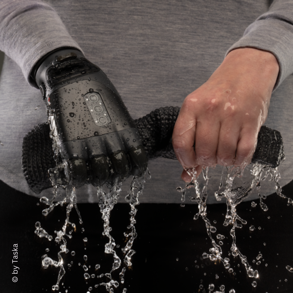 Eine Person wringt einen nassen, schwarzen Schwamm aus. Eine der Hände ist eine moderne, wasserfeste Taska Handprothese, während die andere Hand natürlich ist. Wasser fließt in Strömen aus dem Schwamm und zeigt die Funktionalität und Wasserbeständigkeit der Prothese.